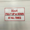 A rectangular aluminum composite sign with a red and white color scheme, text instruction 'DOOR FULLY UP OR DOWN AT ALL TIMES'