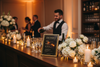 Bar setup with drinks menu, bottles, and floral arrangements in a warm-lit room.