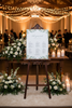 Decorative setup with a seating chart on an easel surrounded by flowers and candles in a warmly lit room.