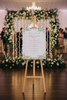 Wedding ceremony setup with floral arch and wooden easel displaying a wedding timeline.